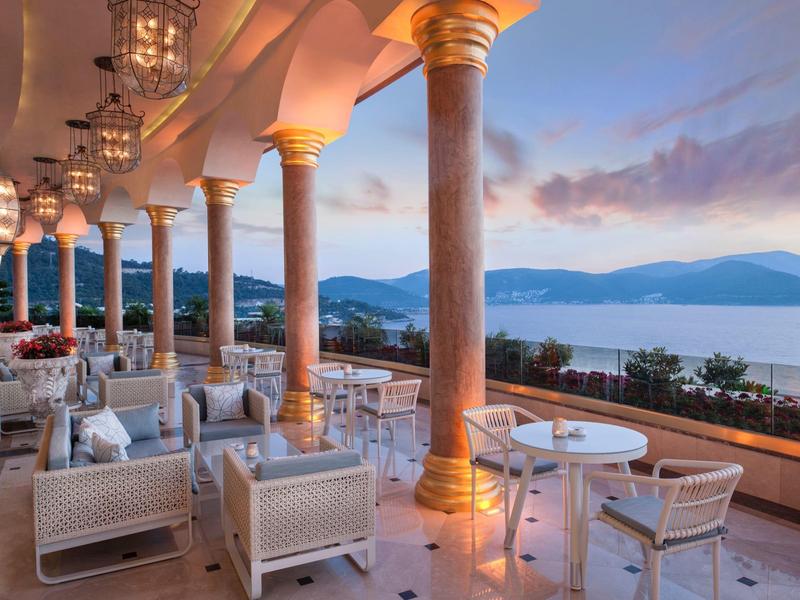 Terrace with seating and columns overlooking mountains and water at sunset.