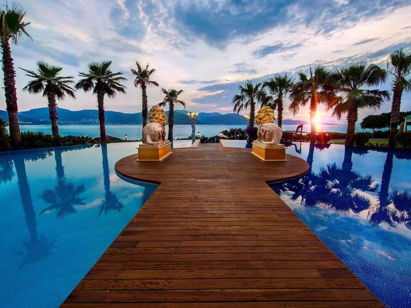 Wooden walkway leads to a pool with palm trees and a sunset view over the sea.