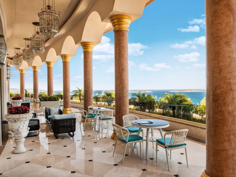 Covered terrace with columns, tables, and chairs overlooking the sea under a clear sky.