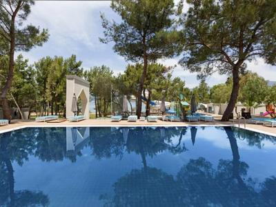 Niebieski basen otoczony drzewami i leżakami pod częściowo zachmurzonym niebem.