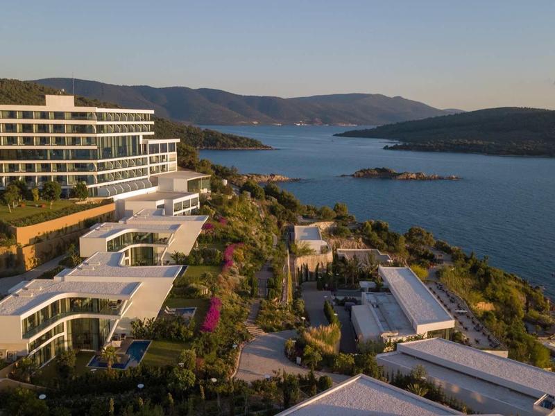 Moderní hotelový komplex na svahu s výhledem na moře a okolní kopce při západu slunce.