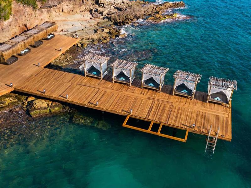 Dřevěné molo s lehátky a slunečníky na průzračném moři u skalnatého pobřeží.