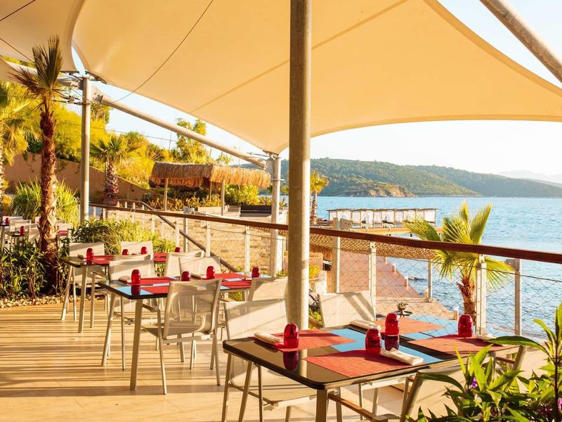 Restaurace pod širým nebem s stoly u moře a výhledem na pobřeží při západu slunce.