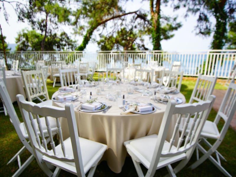 Ronde tafel met witte stoelen op terras met uitzicht op zee en bomen.