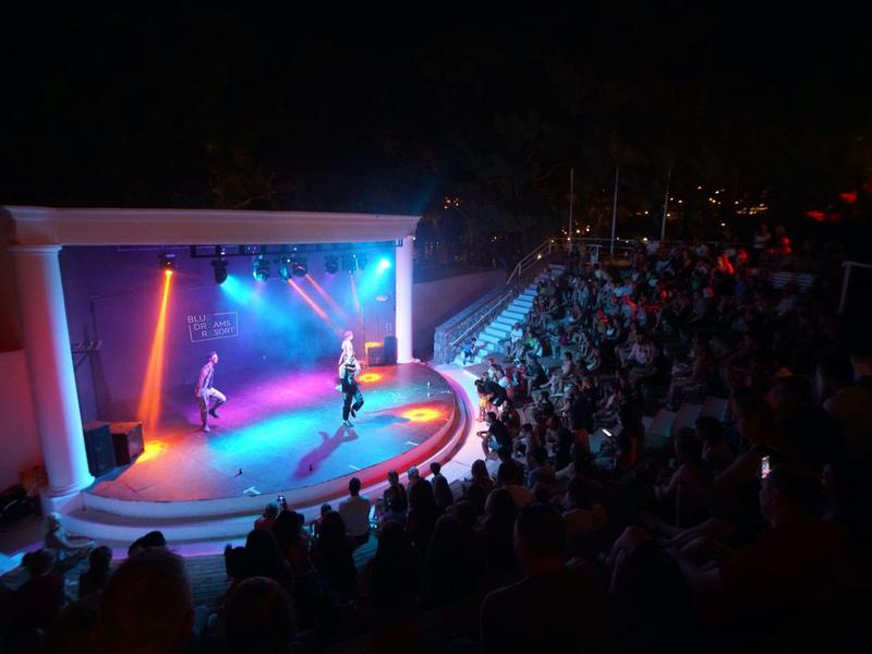 Openluchttheater met artiesten voor groot publiek tijdens avond evenement bij hotel.