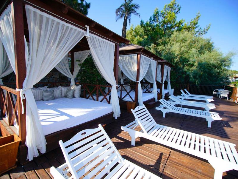 Witte ligstoelen en hemelbedden met gordijnen op een houten terras onder een blauwe lucht.