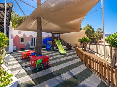 Buitenspeelplaats met glijbanen, kleurrijke tafels en een zonnescherm naast een roze gebouw onder een blauwe hemel.