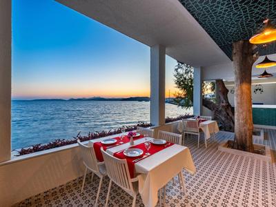 Buiten eetgedeelte met witte tafelkleden aan zee bij zonsondergang, met betegelde vloeren en zuilen.