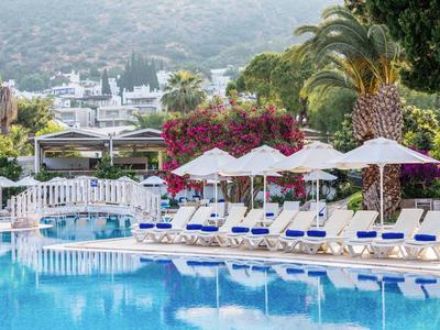 Pool mit Sonnenliegen und weißen Sonnenschirmen, umgeben von Palmen und Berglandschaft.