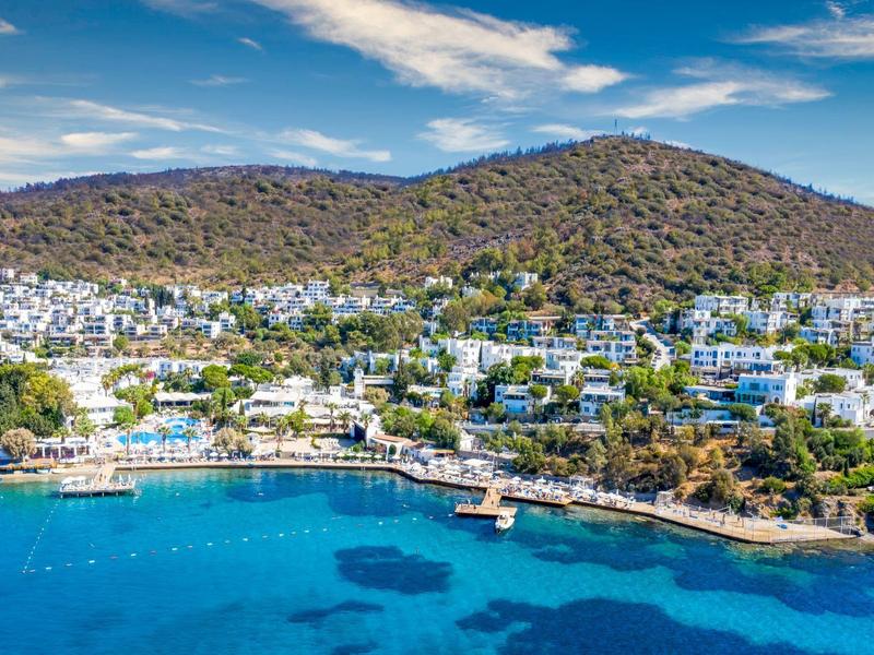 Kuststad met witte gebouwen op heuvels, blauwe zee en een pier onder een partly bewolkte hemel.