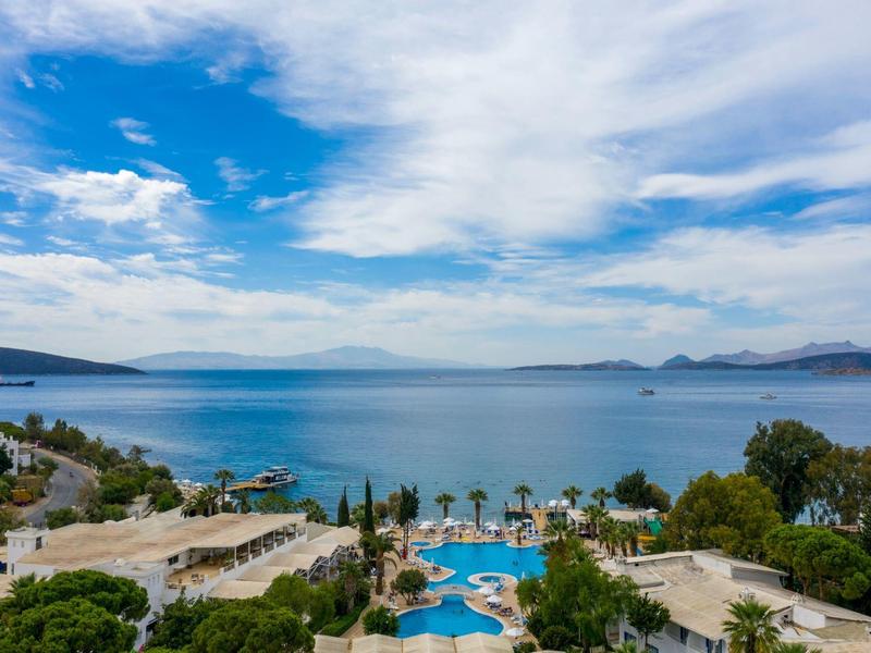 Resort met zwembad en palmbomen aan een uitgestrekte blauwe zee onder een gedeeltelijk bewolkte hemel.