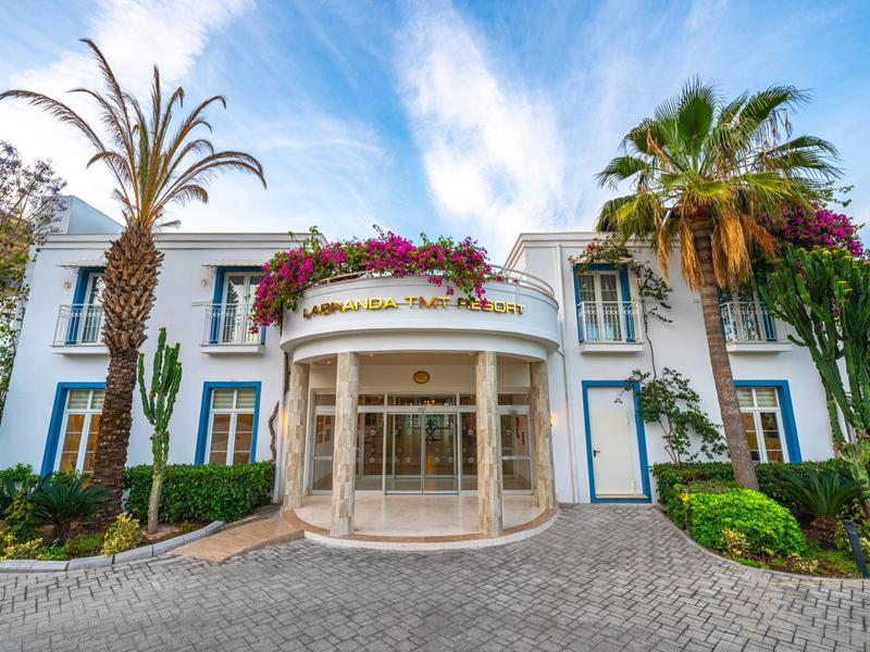 Hotelentree met palmbomen en paarse bloemen onder een blauwe lucht.