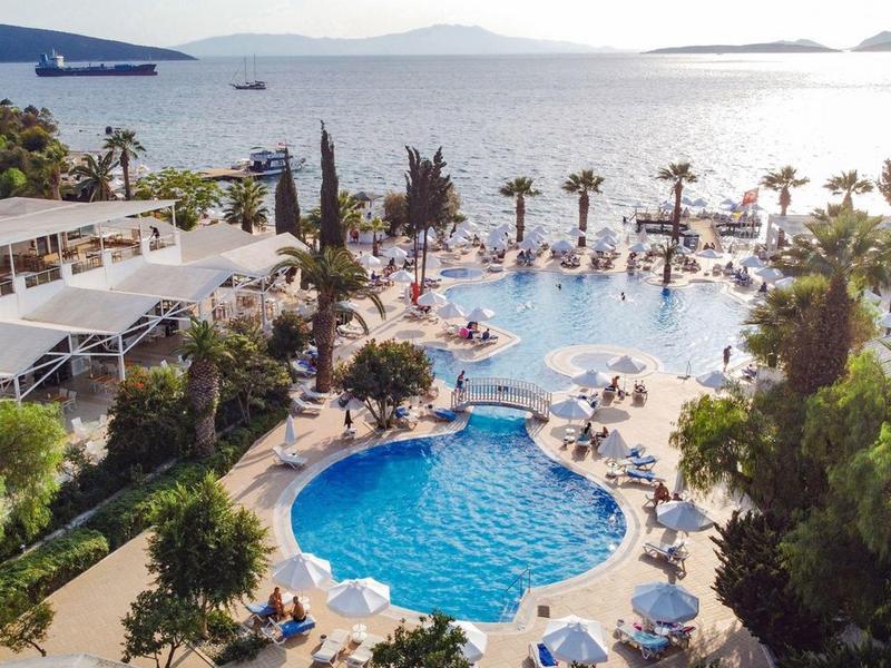 Resort zwembad met rond zwembad, ligstoelen, palmbomen en uitzicht op zee bij zonsondergang.