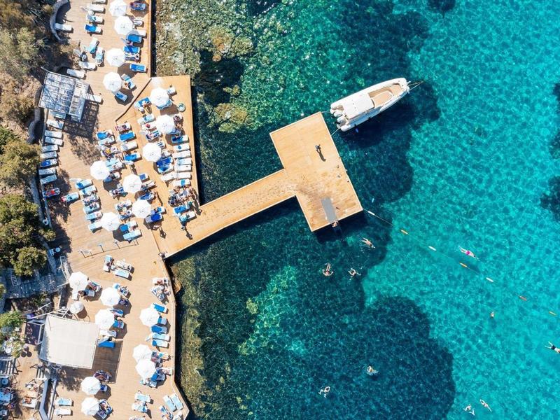 Luchtfoto van een aanlegsteiger met ligstoelen en parasols langs de rotsachtige kust bij helder blauw water.