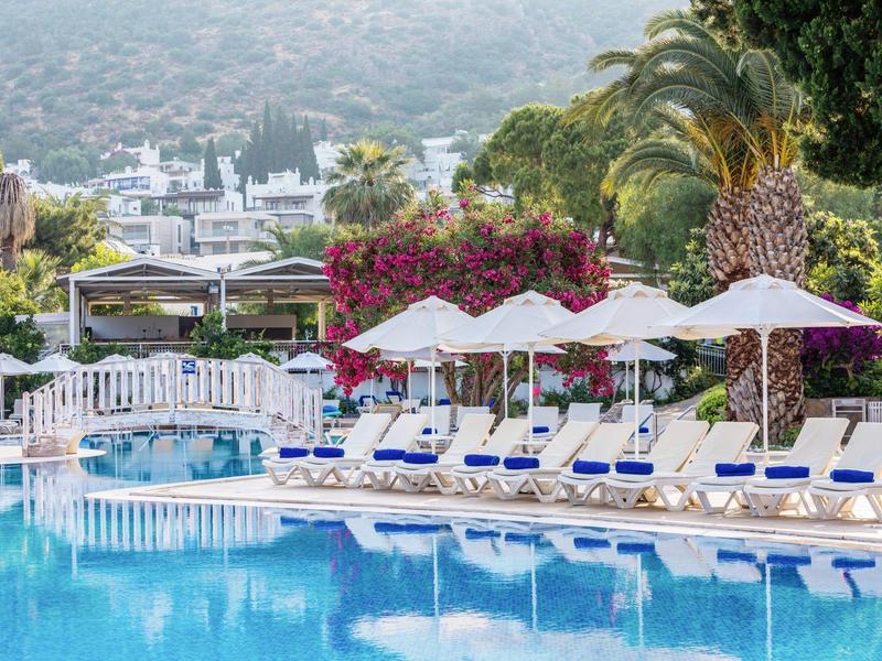 Resortzwembad met ligstoelen, witte parasols, palmbomen en bergachtergrond.