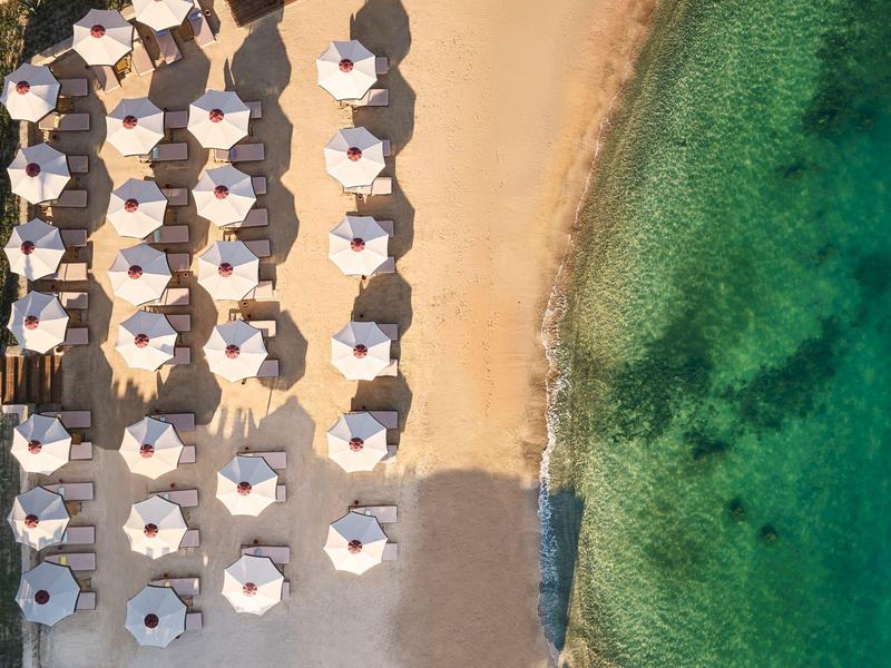 Eine Strandlandschaft mit Reihen weißer Sonnenschirme auf goldenem Sand neben klarem, grünlichem Meer.