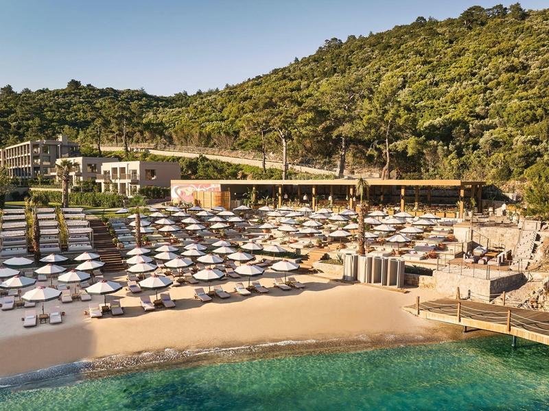 Strand mit weißen Sonnenschirmen, Sand, türkisfarbenem Wasser und grünen bewaldeten Hügeln im Hintergrund.