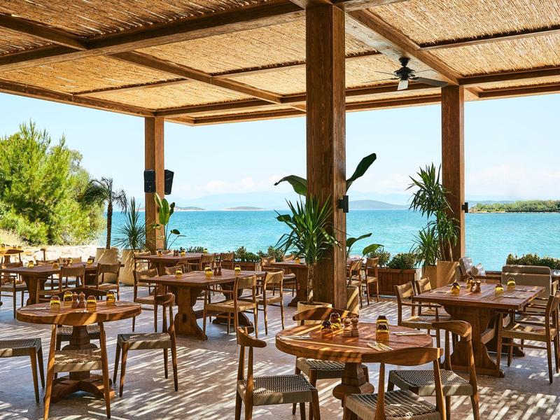 Offenes Restaurant mit Holztischen und Stühlen, Blick auf türkisfarbenes Meer und grüne Pflanzen.