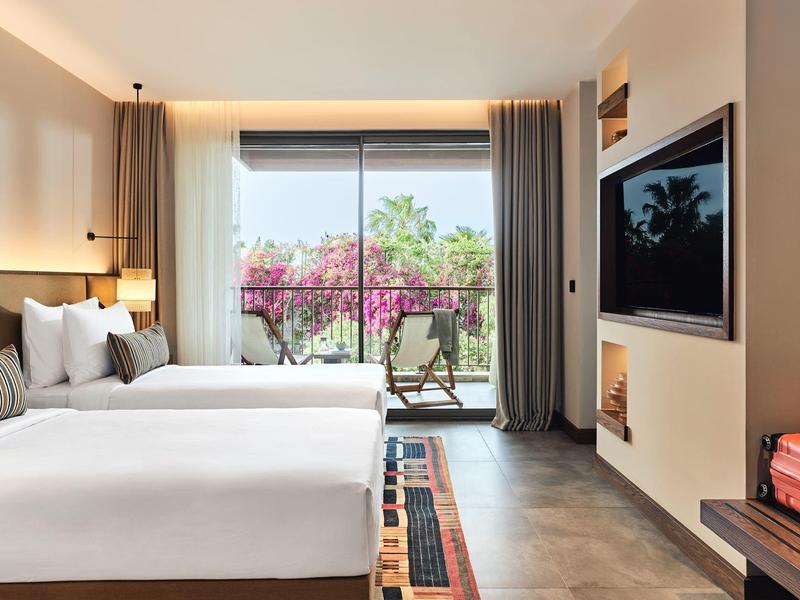 Hell eingerichtetes Schlafzimmer mit zwei großen Betten und Balkon mit Blick auf grüne Pflanzen.