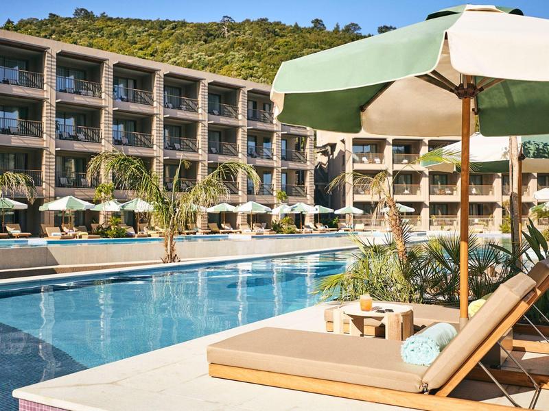 Moderner Hotelpool mit Liegestühlen, Sonnenschirmen und grünen Hügeln im Hintergrund.