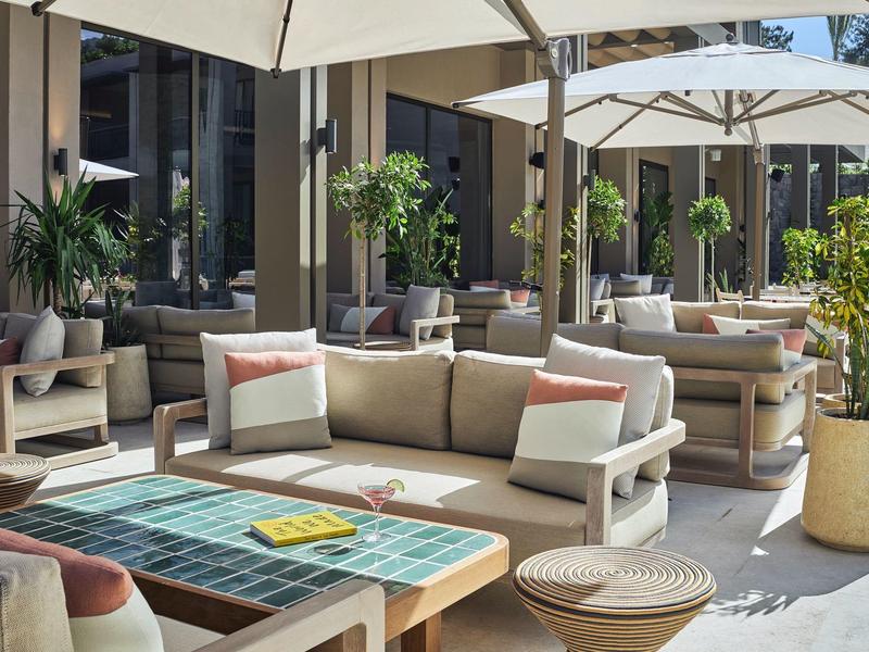Terrasse mit bequemen Sofas, Tischen und weißen Sonnenschirmen, umgeben von Pflanzen.