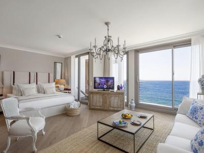 Camera d'hotel ben arredata con vista sul mare, letto, divano e tavolo in vetro in uno spazio luminoso.