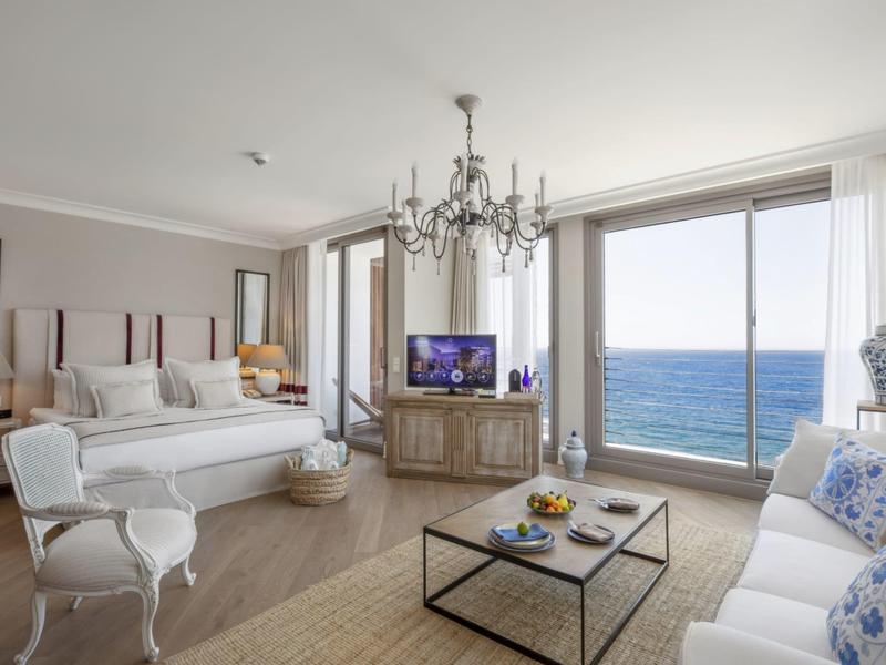 Camera d'hotel ben arredata con vista sul mare, letto, divano e tavolo in vetro in uno spazio luminoso.