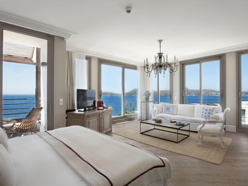 Camera d'hotel luminosa con vista sul mare, letto, divano, TV e balcone con posti a sedere.