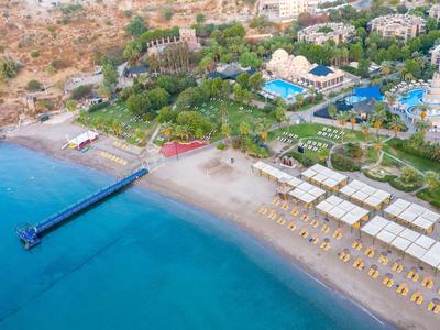 Aerial view of a resort with a beach, sun umbrellas, and a pier in clear blue water.