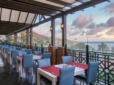 Covered hotel terrace with blue chairs, white tables, and sea view at sunset.
