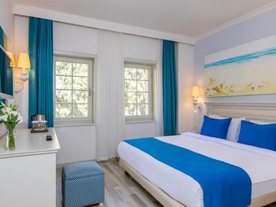 Bright hotel room with double bed, blue and white bedding, windows with blue curtains, and a desk.
