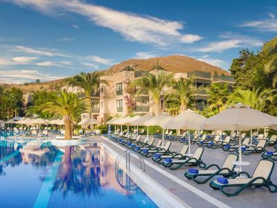 Großer Pool mit blauer Wasserfläche, Liegestühlen und Sonnenschirmen vor einem Hotelgebäude.