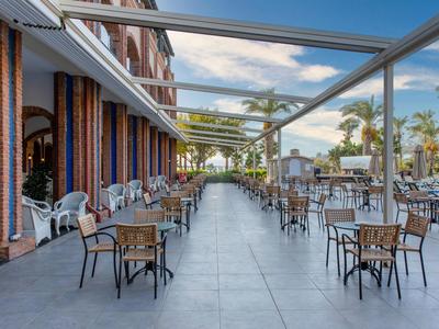 Large covered terrace with tables and chairs beside a hotel, surrounded by palm trees.