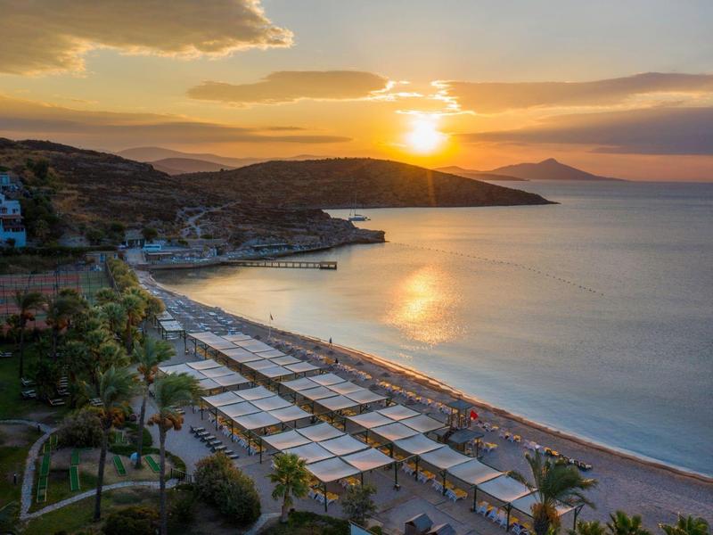Sunset over a beach with loungers and shaded cabanas beside a calm sea.