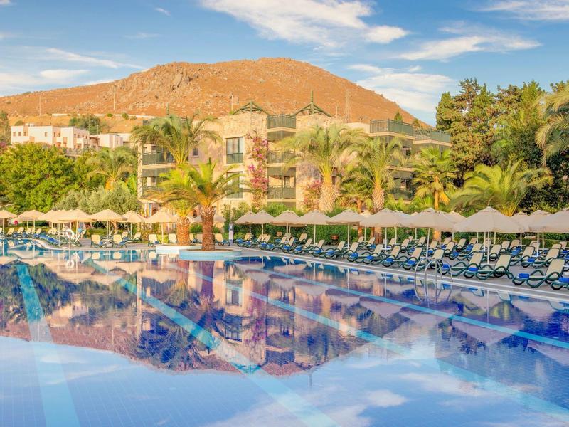Large pool with lounge chairs and umbrellas in front of a hotel complex and mountains in the background.