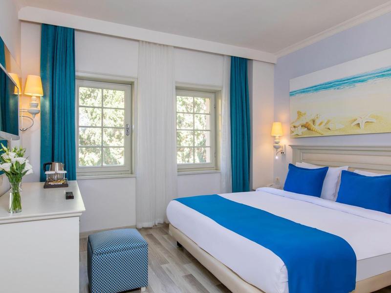 Bright hotel room with double bed, blue and white bedding, windows with blue curtains, and a desk.