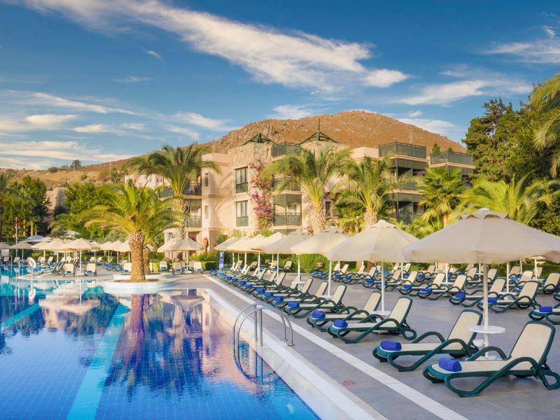 Hotel complex with swimming pool, sun loungers, and palm trees under a blue sky.