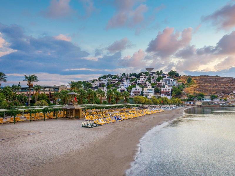Beach with sun loungers and white houses on a hilly coastal town under a cloudy sky.