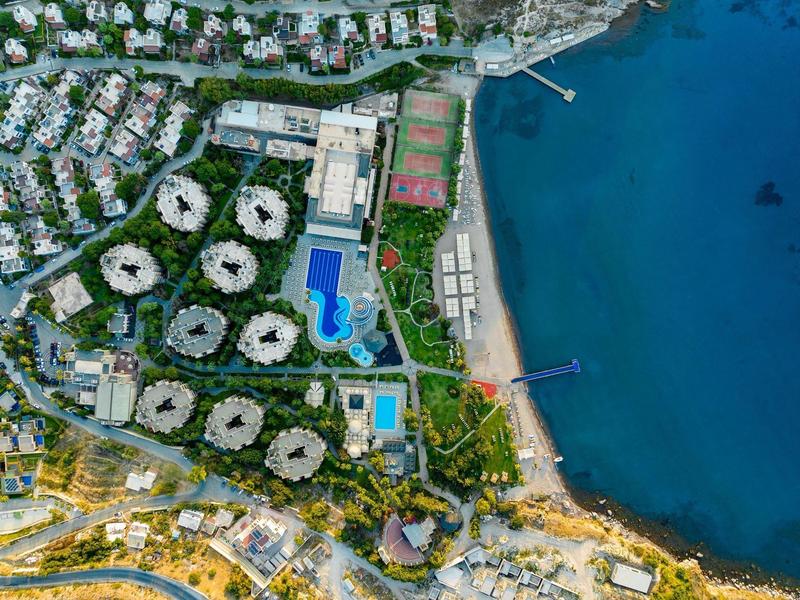 Aerial view of a coastal resort with circular buildings, pools, and tennis courts by the sea.