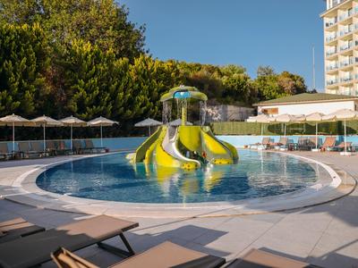 Round pool with multiple water slides and lounge chairs in front of a hotel building.