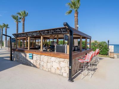 Offener Strandbar mit Holzstruktur, Palmen und Sitzplätzen am Meer unter blauem Himmel.