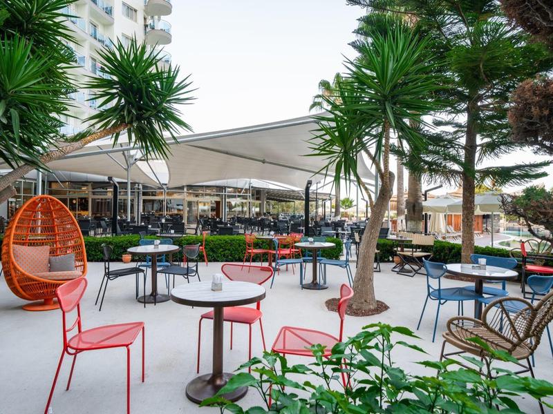 Taras na świeżym powietrzu z kolorowymi stołami, krzesłami i drzewami, relaksująca atmosfera.