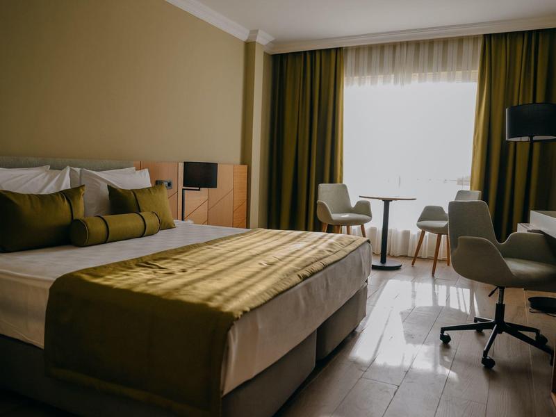 Pokój hotelowy z łóżkiem podwójnym, biurkiem, dwoma krzesłami i dużym jasnym oknem.