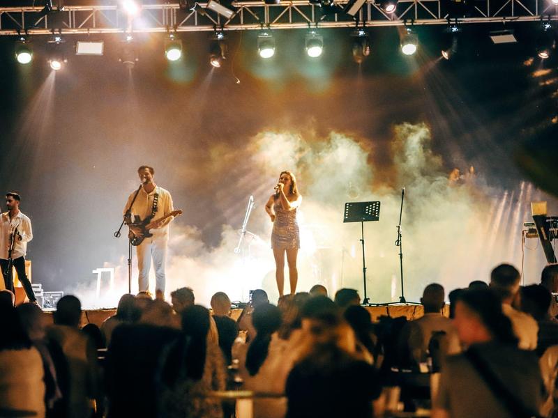 Koncert na żywo z trzema muzykami na scenie przed entuzjastyczną publicznością pod kolorowymi światłami.