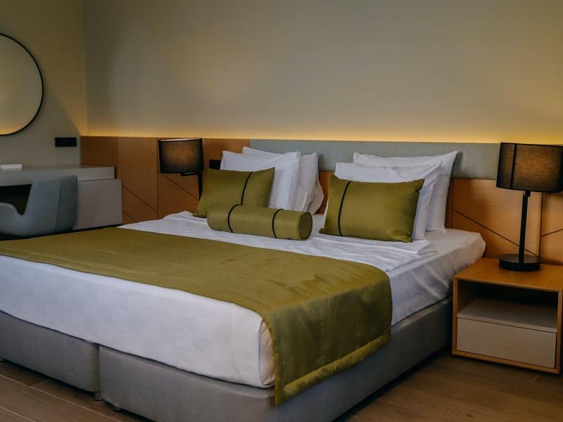 Nowoczesny pokój hotelowy z podwójnym łóżkiem, zielonymi poduszkami i lampką na stoliku nocnym.