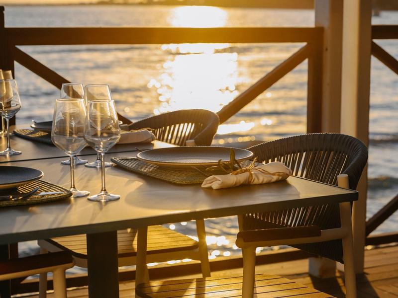 Drewniany stół nakryty szkłem i talerzami nad wodą o zachodzie słońca.