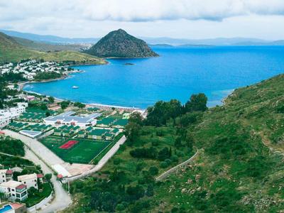 Pobřežní krajina s zálivem, horami, domy a zelenými poli pod zataženou oblohou.