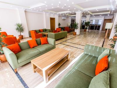 Světlá hotelová hala se zelenými pohovkami, oranžovými polštáři a květinami v květináčích.