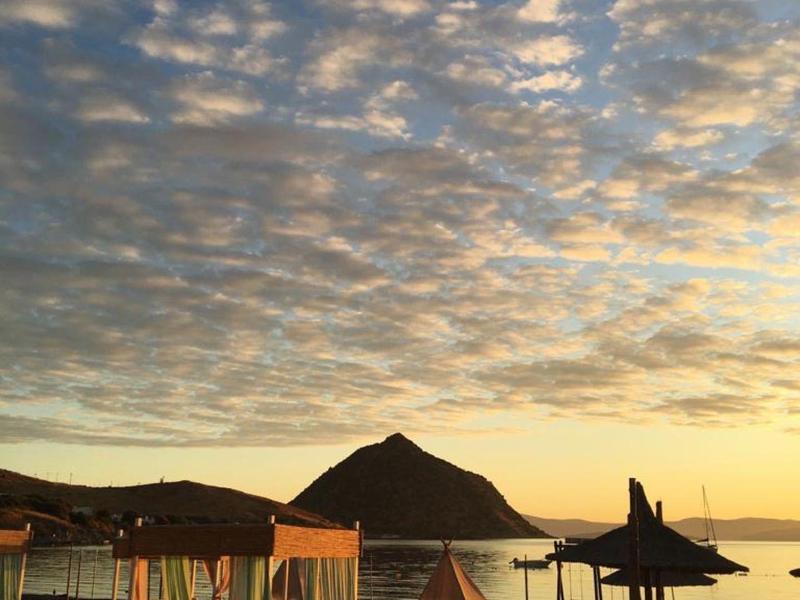 Západ slunce nad klidnou vodou s chatkami a lehátky v popředí.