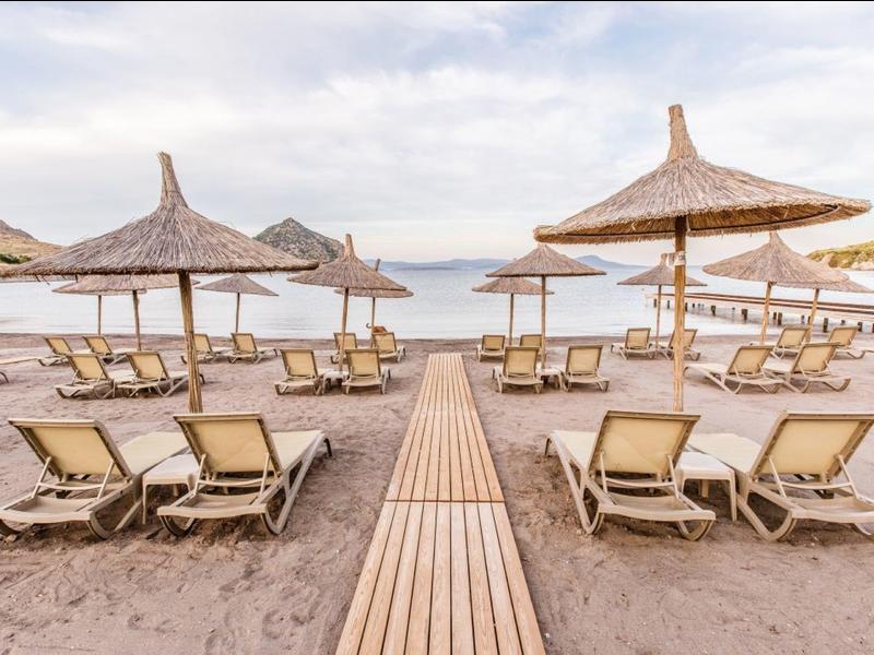 Písčitá pláž s lehátky a slunečníky vedle dřevěné cesty pod oblačnou oblohou.